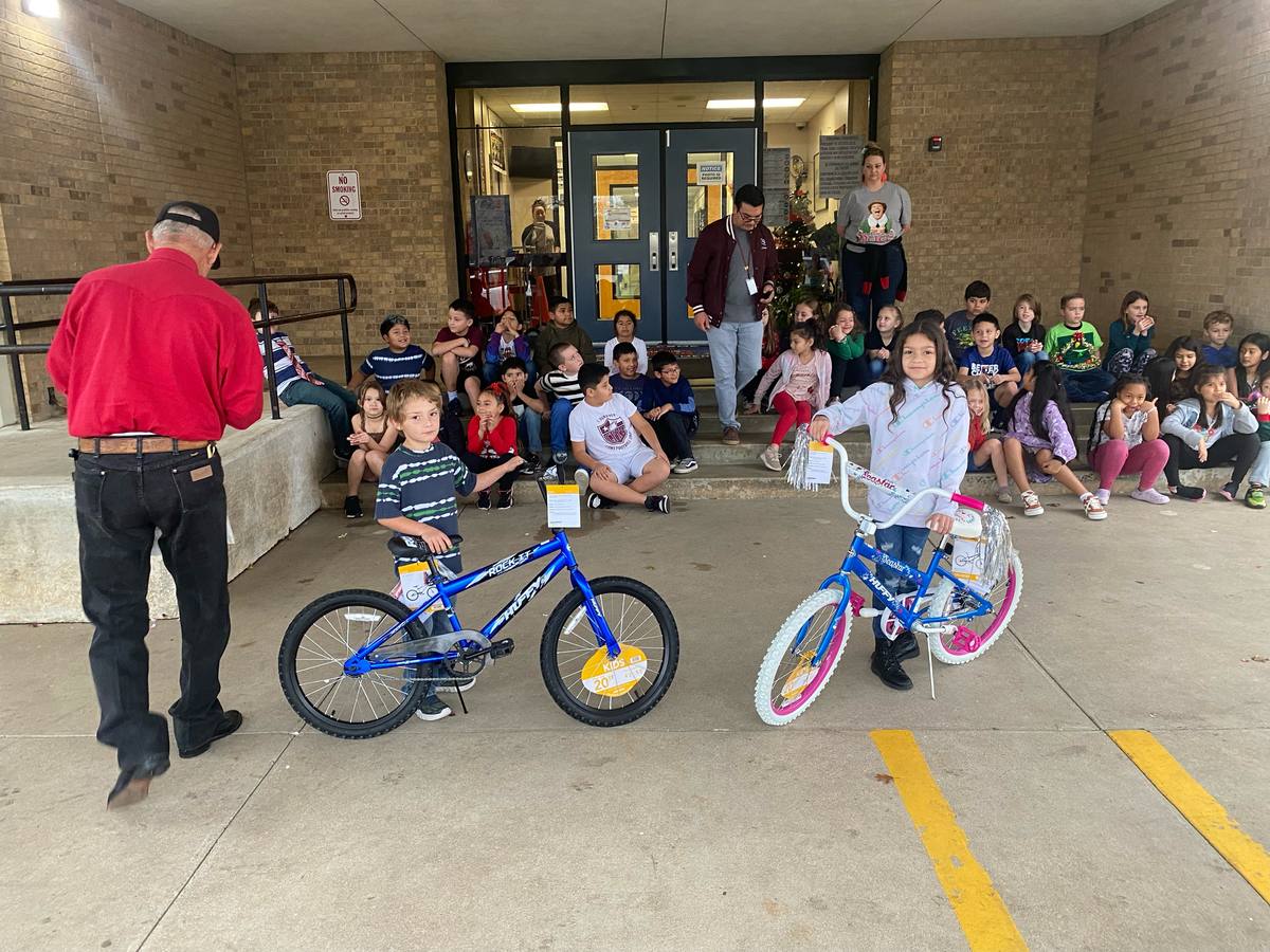 Two students stand with their new bicycles outside a school entrance while classmates watch from the steps